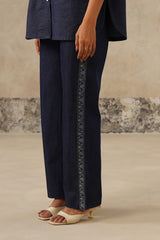 A person wearing dark navy wide-leg pants with a decorative vertical embroidered stripe down the side, paired with a matching navy top and cream heeled sandals, standing against a textured beige wall.