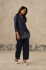 A woman modeling a navy blue two-piece outfit consisting of a long-sleeve tunic top with floral embroidery and matching wide-leg trousers, set against a textured beige studio backdrop. denim coord set, cutwork embroidery