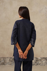 A woman viewed from behind wearing a dark navy blue kurta with three-quarter sleeves and white embroidered details on the cuffs, paired with matching navy trousers, set against a textured beige wall.