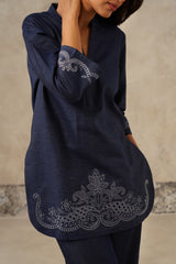 A woman wearing a dark navy denim blue tunic top with intricate silver-gray embroidery on the sleeve and hem, paired with matching navy pants, set against a textured beige wall.