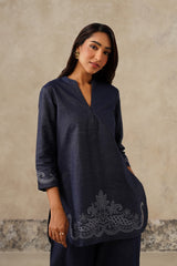 A woman modeling a navy blue denim tunic top with tonal cutwork embroidered detailing on the cuffs and hem, set against a textured beige wall.
