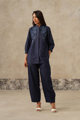 A woman modeling a denim coord set consisting of a long-sleeve button-up top with floral embroidery on the chest and matching wide-leg cropped pants, set against a textured beige studio backdrop.