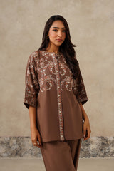 A woman modeling a brown three-quarter sleeve kurta with intricate white and beige paisley embroidery, paired with matching brown trousers, set against a textured beige wall.