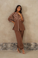 A woman models a brown two-piece outfit consisting of a short-sleeved button-up top with beige paisley embroidery and matching wide-leg trousers, standing against a textured beige wall.