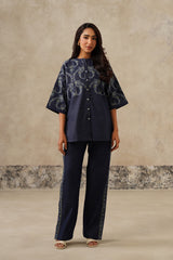 A woman modeling a navy blue two-piece set consisting of a short-sleeved button-up top with a white floral pattern and matching navy wide-leg trousers with lace trim, set against a textured beige studio backdrop.