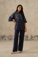 A woman modeling a denim coord set consisting of a short-sleeved button-up top with white paisley embroidery and matching wide-leg trousers with embroidered side details, set against a textured beige studio backdrop.