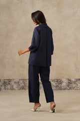 A woman is shown from the back wearing a dark navy blue two-piece set consisting of a long-sleeve button-up shirt and matching wide-leg trousers, paired with white high-heeled sandals, set against a textured beige studio backdrop.