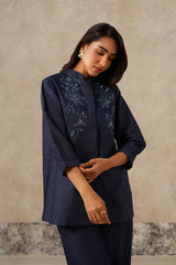 A woman wearing a dark navy blue embroidered kurta with matching trousers, standing against a textured beige wall.