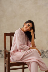 A woman wearing a light pink traditional outfit sits on a dark wooden chair against a textured beige wall.